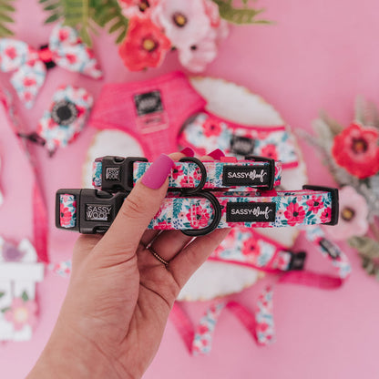 A hand with pink nail polish holds two Sassy Woof "Dog Collar - Floral Frenzy" collars, featuring floral patterns, with matching pet accessories and vibrant flowers on a pink background.
