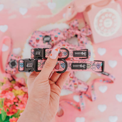 A hand with heart nail art holds two Sassy Woof "I Chews You" pink adjustable dog collars, decorated with colorful hearts and the brand logo. The background shows flowers, a pink rotary phone, and heart patterns.
