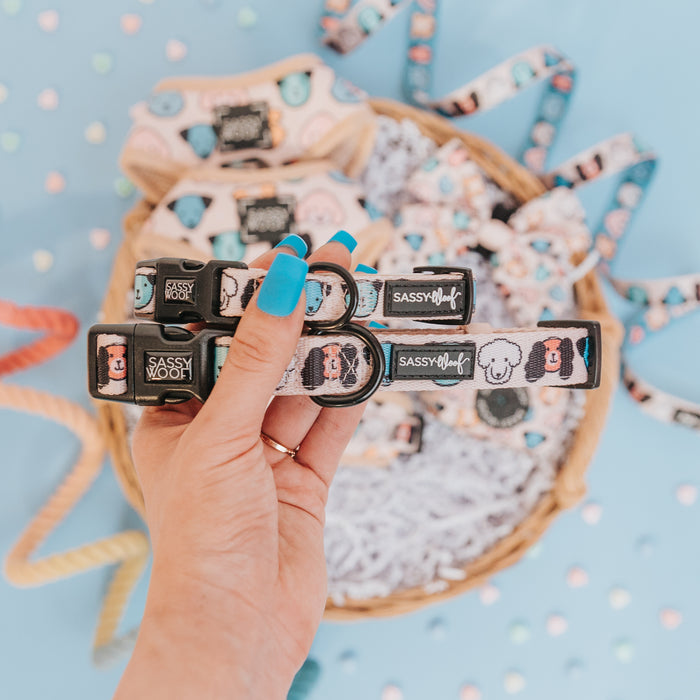 A hand with blue-painted nails holds two pastel Sassy Woof Dog Collar - Mixed With Love collars, featuring cartoon dog faces, above a basket of similar collars on a light blue heart-decorated background.