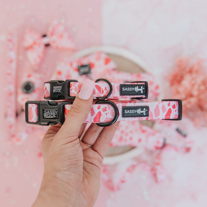 A hand with pink painted nails holds two Sassy Woof "Dog Collar - Mykonos" collars featuring pink and red designs. Matching pet accessories are displayed on a pink surface in the background.
