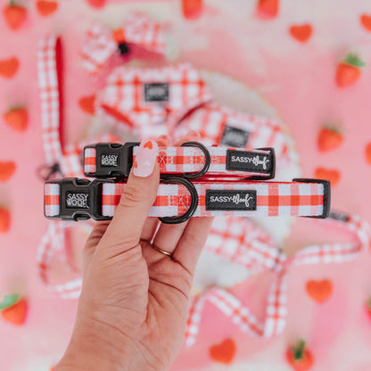 A hand with strawberry nail art holds two Sassy Woof "Picnic Fur Two" dog collars, each with a sturdy D-ring. The pink background features scattered strawberries and additional checkered neoprene collars.