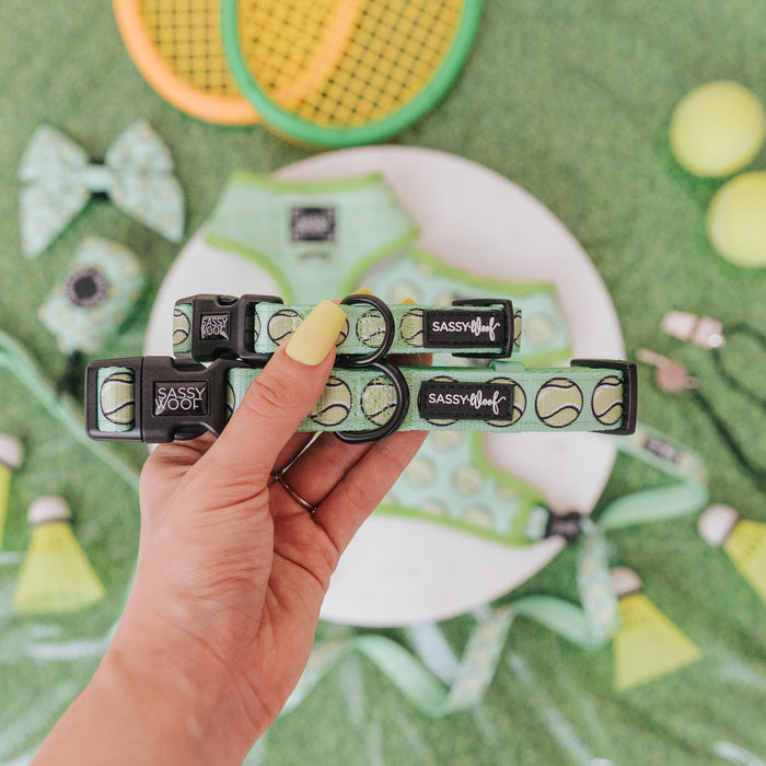 A hand with yellow nails holds two Sassy Woof "Serving Up Sass" dog collars. In the background are green dog accessories, tennis rackets, balls, and shuttlecocks on grass. These adjustable collars are perfect for sporty pups!.