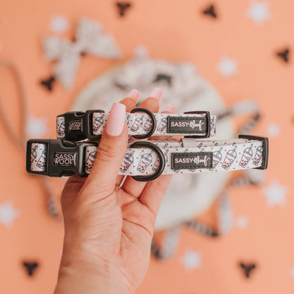 A hand with long, manicured nails holds two Sassy Woof "Spill the Tea" dog collars, featuring black brand labels, fun illustrated patterns, and sturdy D-rings, set against a peach background with scattered bone shapes.