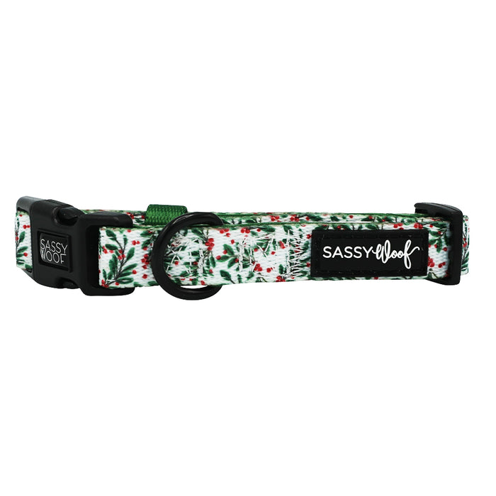 The Sassy Woof Dog Collar - Under the Mistletoe features a green and red foliage pattern, black plastic buckles, a metal ring, and an adjustable fit with a black SASSY WOOF tag—perfect to pair with matching accessories.