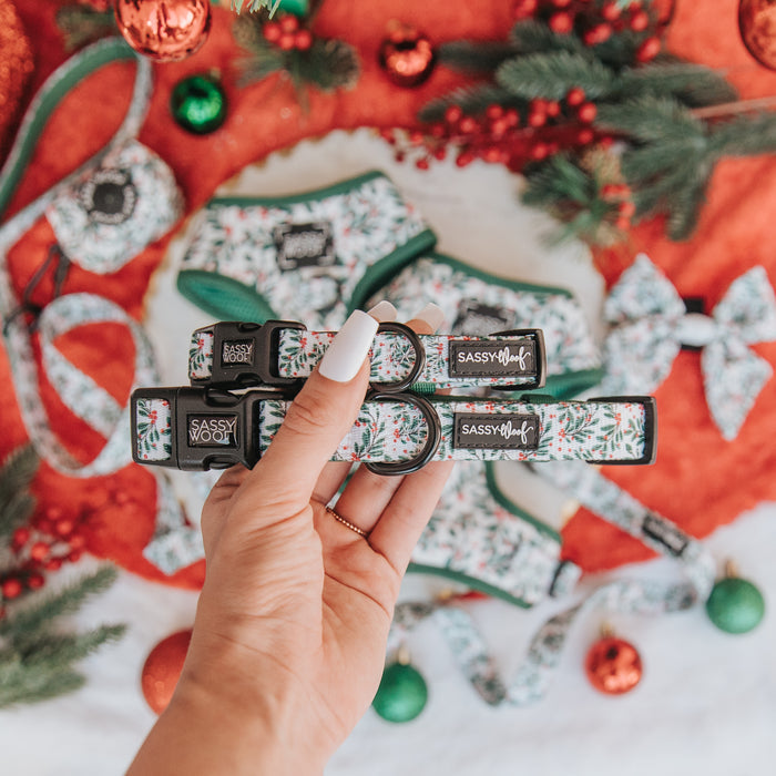 A hand with white-painted nails holds two "Under the Mistletoe" adjustable dog collars by Sassy Woof. The background has matching accessories, pine branches, and Christmas ornaments on a red and white surface.