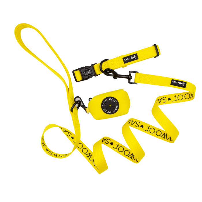 The Sassy Woof Dog Collar Three Piece Bundle in Neon Yellow features a vibrant dog leash, collar, and waste bag holder. The leash and collar display bold black "SASSY WOOF" text, while the waste bag holder is adorned with a round black logo. All items are arranged neatly on a white background.