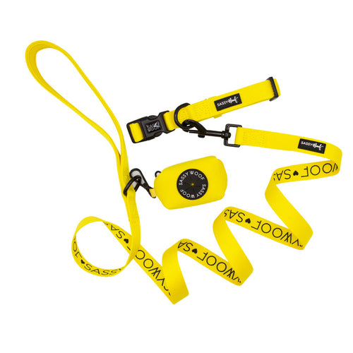 The Sassy Woof Dog Collar Three Piece Bundle in Neon Yellow features a vibrant dog leash, collar, and waste bag holder. The leash and collar display bold black "SASSY WOOF" text, while the waste bag holder is adorned with a round black logo. All items are arranged neatly on a white background.