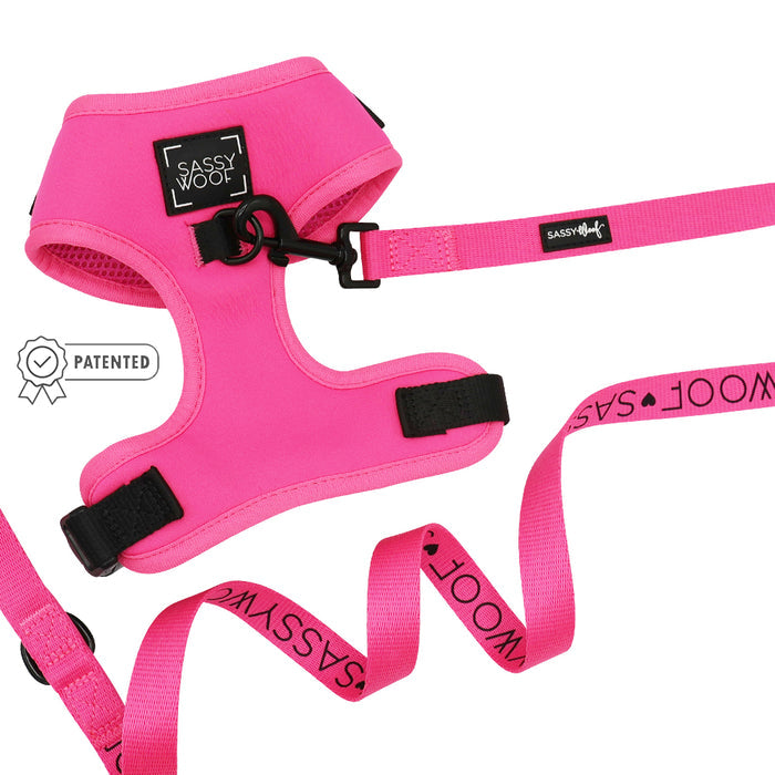 The Sassy Woof Dog Two Piece Bundle - Neon Pink is displayed on a white background. This adjustable harness has a black buckle, and the matching leash features repeated SASSY WOOF branding. A patented badge is also visible.