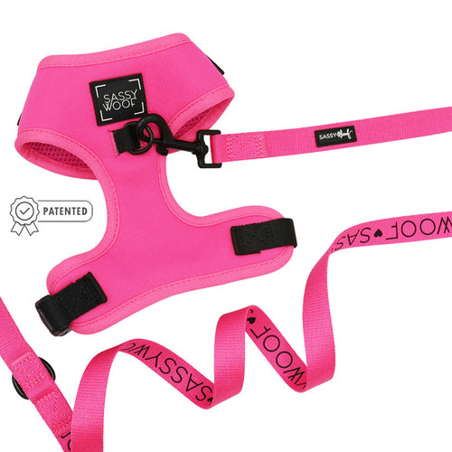 The Sassy Woof Dog Two Piece Bundle - Neon Pink is displayed on a white background. This adjustable harness has a black buckle, and the matching leash features repeated SASSY WOOF branding. A patented badge is also visible.