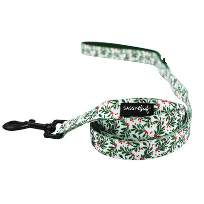 The Sassy Woof Dog Two Piece Bundle - Under the Mistletoe features a green and red holly leash with a black metal clip and branded handle, paired perfectly with an adjustable harness for growing puppies. Shown loosely coiled on white.