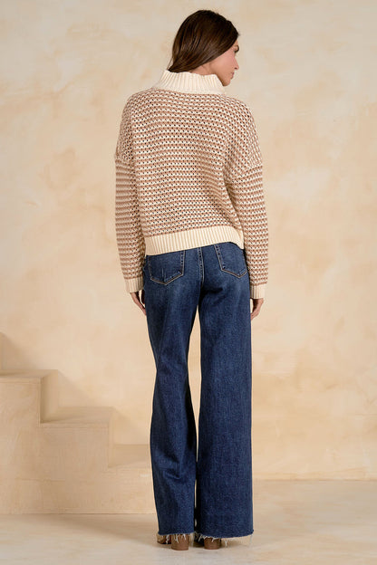 Woman wearing a patterned sweater and blue jeans against a beige wall.