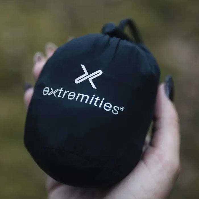 Black stuff bag holding a pair of extremities Paradox lightweight waterproof gloves held in a  persons palm against a blurred natural background