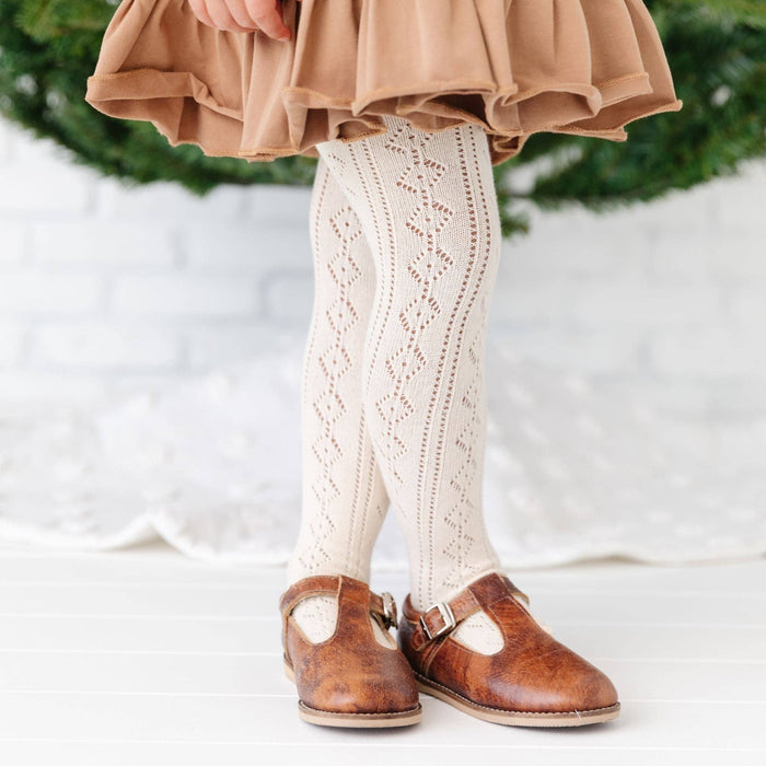 A young child wearing Little Stocking Co. Fancy Knit Tights in Vanilla stands on a white floor before greenery and a white brick wall.