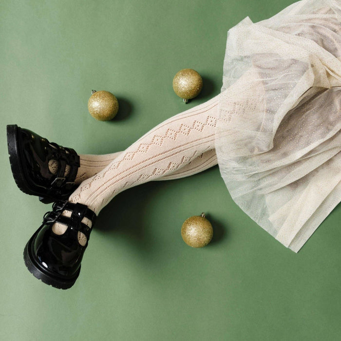 A person wearing Little Stocking Co. Fancy Knit Tights in Vanilla lies on a green surface with four gold Christmas ornaments nearby, paired with black patent shoes and a sparkly tulle skirt.