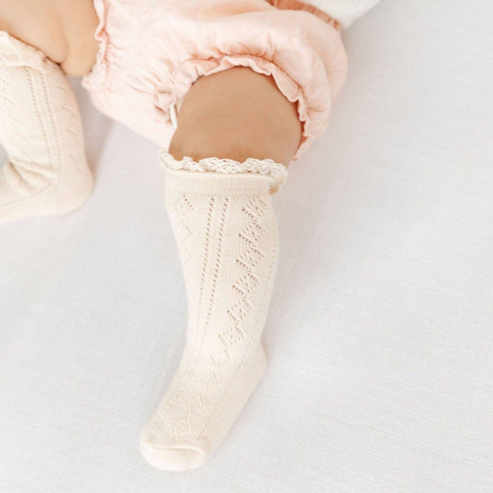 A baby wearing Little Stocking Co. Fancy Lace-Top Knee-High Socks in Vanilla and pale pink ruffled shorts lies on a white blanket.
