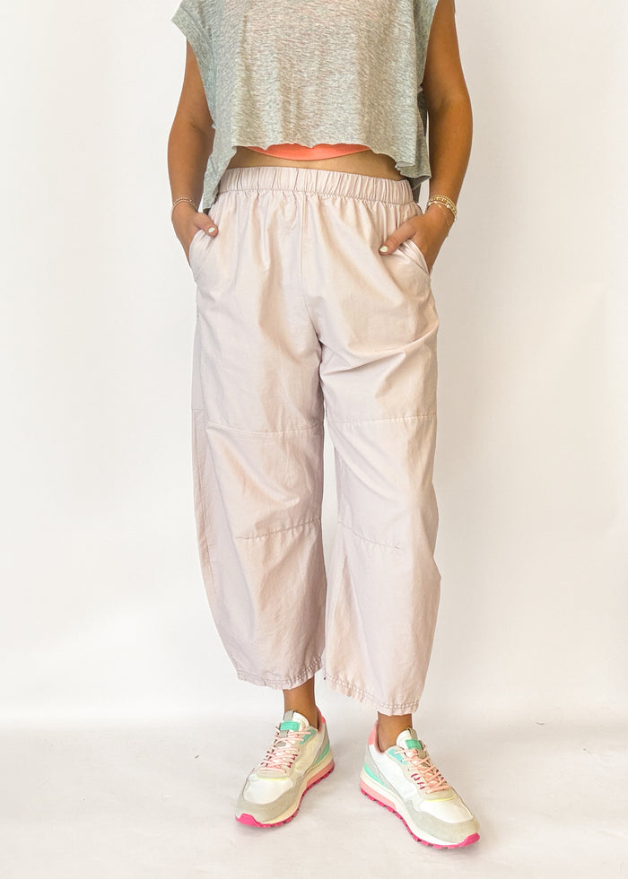 A person wearing loose, light blush-beige cropped pants with an elastic waistband and side pockets, paired with a gray sleeveless crop top and pastel sneakers, standing against a plain light background.