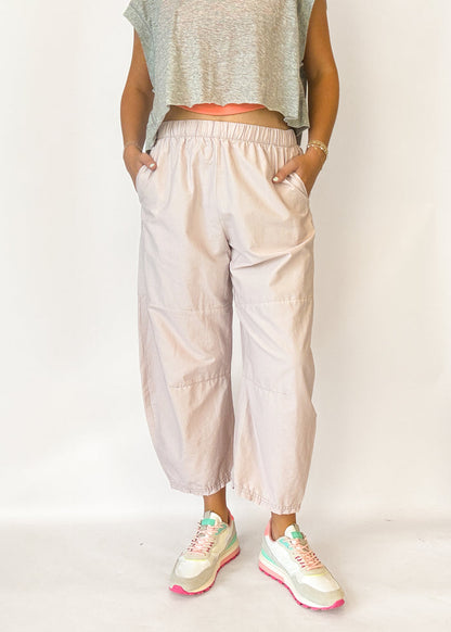 A person wearing loose, light blush-beige cropped pants with an elastic waistband and side pockets, paired with a gray sleeveless crop top and pastel sneakers, standing against a plain light background.