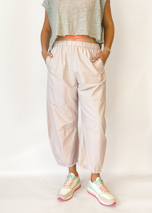 A person wearing loose, light blush-beige cropped pants with an elastic waistband and side pockets, paired with a gray sleeveless crop top and pastel sneakers, standing against a plain light background.