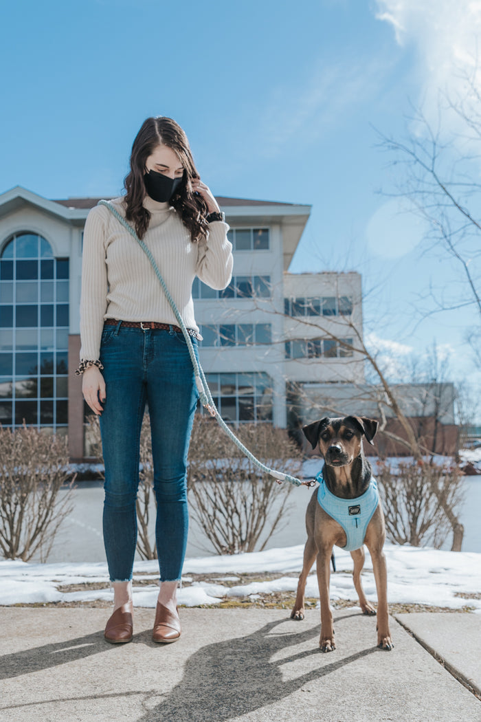 A woman wearing a mask stands outside on a sidewalk, holding the Sassy Woof Hands-Free Rope Leash - Azul attached to a dog in a blue harness. An office building and leafless bushes are visible in the background beneath a blue sky.