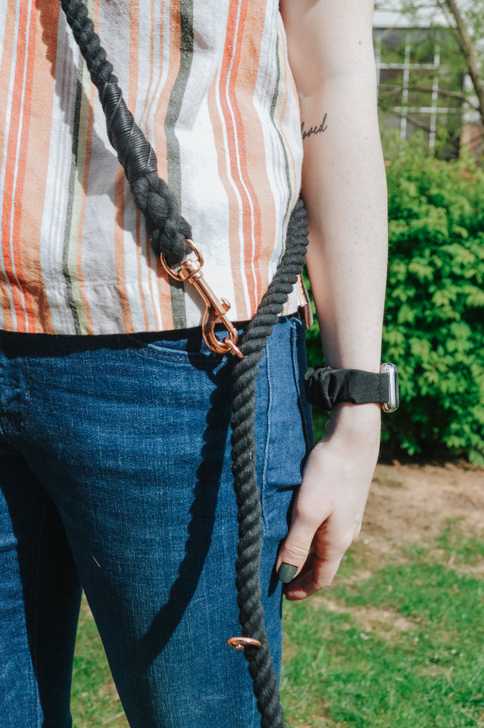 A person wearing blue jeans, a striped shirt, and a black watch holds the Sassy Woof Hands-Free Rope Leash - Noir outdoors, with greenery in the background.