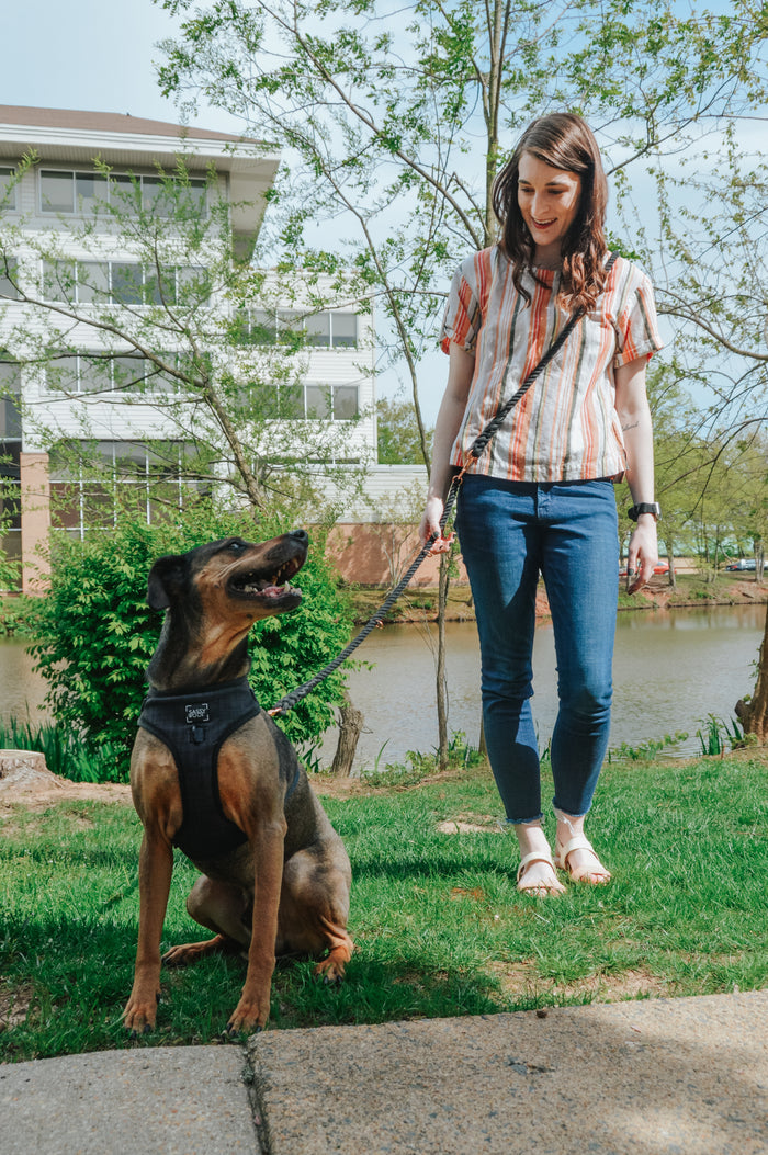 A woman in a striped shirt and jeans smiles while holding the leash of a large, seated dog outside by a pond and trees on a sunny day. The dog sits patiently at her side, secured with the Sassy Woof Hands-Free Rope Leash - Noir, which adds both style and function to their happy outing.
