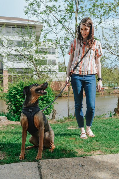A woman in a striped shirt and jeans smiles while holding the leash of a large, seated dog outside by a pond and trees on a sunny day. The dog sits patiently at her side, secured with the Sassy Woof Hands-Free Rope Leash - Noir, which adds both style and function to their happy outing.