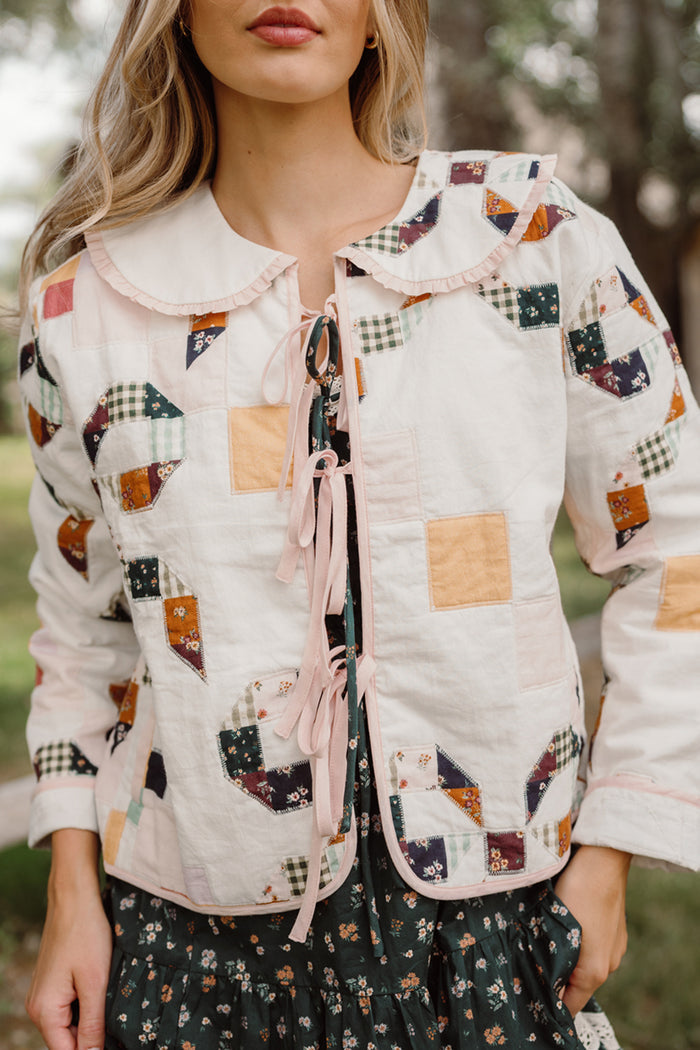 A woman wears the Ivy City Co Harper Jacket in Quilted Bow, a white quilted cotton jacket adorned with colorful patchwork patterns and featuring a large collar, paired with a dark floral dress. She stands outdoors in a natural setting.