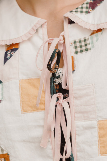 Close-up of the Ivy City Co Harper Jacket in Quilted Bow, a women’s cotton jacket featuring colorful fabric patches, a pink ruffle collar, and pink ribbon lace-up closure, styled over a dark floral top.