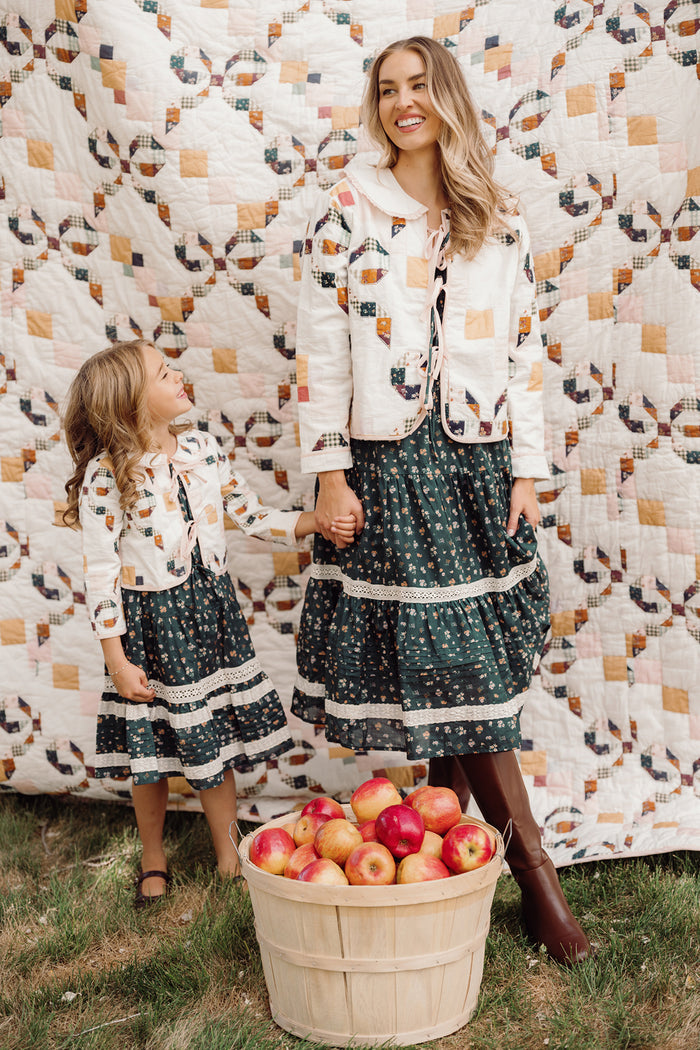 A woman and a young girl, both dressed in matching Ivy City Co Harper Jackets in Quilted Bow, stand hand in hand and smiling before a quilt, with a basket of red apples resting on the grass nearby.