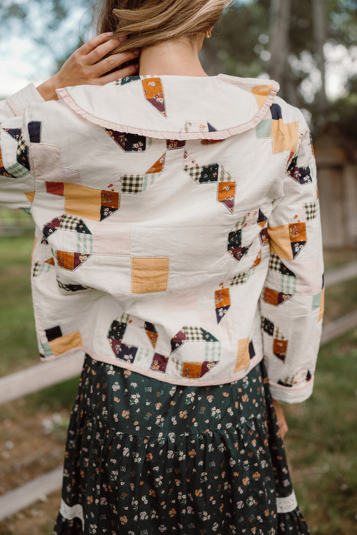 An individual wearing the Ivy City Co Harper Jacket in Quilted Bow—a women’s cotton jacket with a ruffled collar and assorted patterned fabric patches—stands outdoors. Shown from the back, they pair the jacket with a dark floral skirt.
