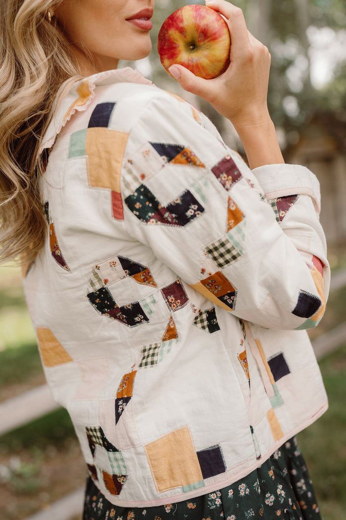 A woman with wavy blonde hair holds an apple near her mouth while wearing the Ivy City Co Harper Jacket in Quilted Bow—a colorful women’s cotton jacket featuring a vibrant patchwork design. She stands outdoors, surrounded by greenery.