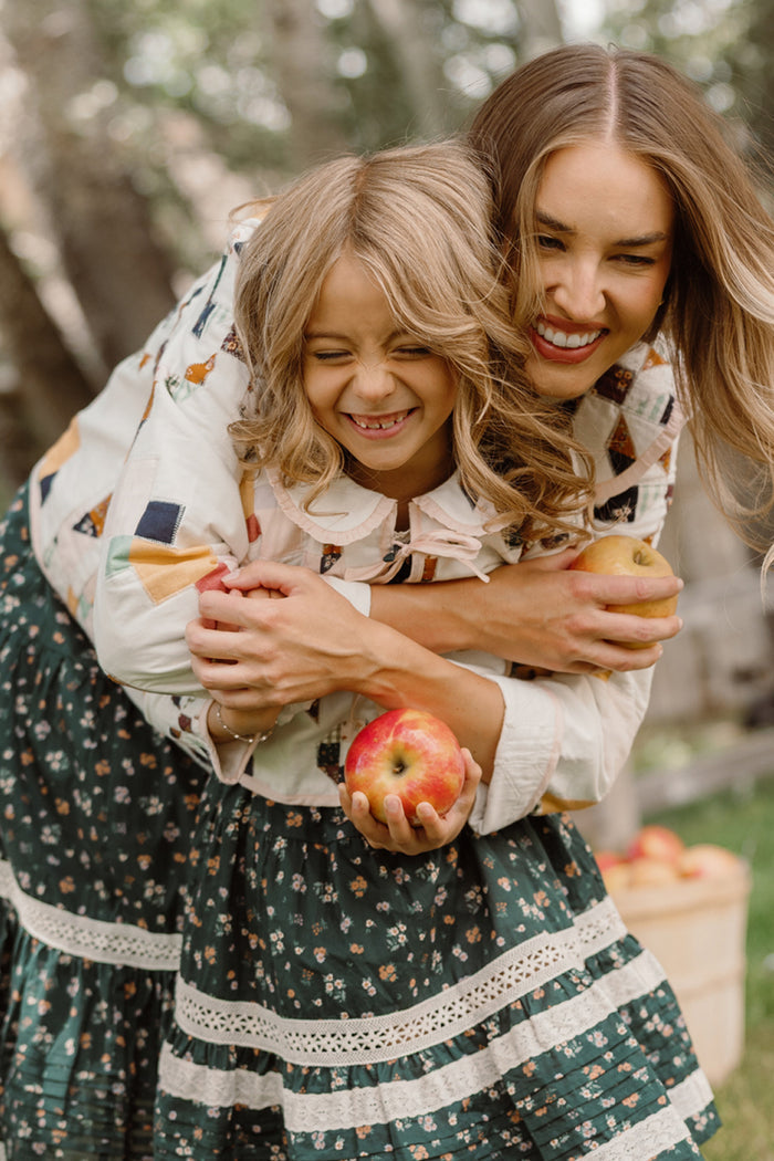 A smiling woman and girl, both in patterned skirts, hug each other while holding apples. The woman is wearing the Harper Jacket in Quilted Bow by Ivy City Co, adding a touch of style to their cheerful moment. They appear happy, standing outdoors with trees and a basket of apples in the background.