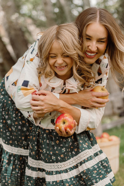 A smiling woman and girl, both in patterned skirts, hug each other while holding apples. The woman is wearing the Harper Jacket in Quilted Bow by Ivy City Co, adding a touch of style to their cheerful moment. They appear happy, standing outdoors with trees and a basket of apples in the background.