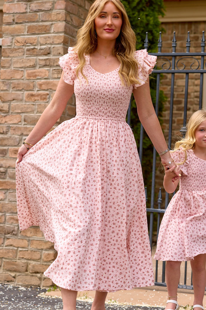 A woman in the Ivy City Co Hattie Dress in Madeline Pink holds hands with a young girl in a matching birthday dress. Both wear charming pink floral cotton midi dresses as they stand by a brick wall and iron gate.