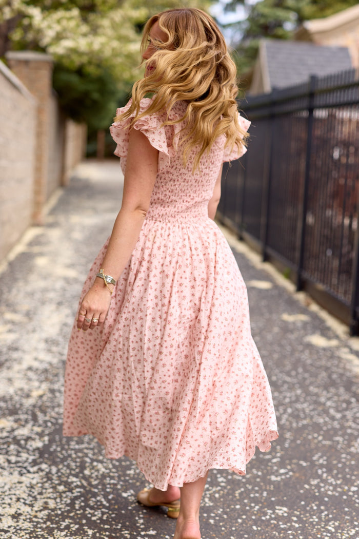A woman with wavy blonde hair walks along a petal-strewn path in the Ivy City Co Hattie Dress in Madeline Pink, her back turned and head slightly to the side, wearing sandals.