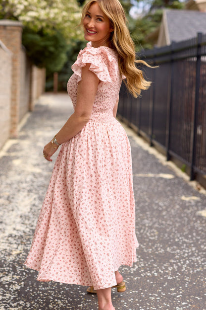 A woman with long, wavy hair smiles while turning on a tree-lined path. She wears the Ivy City Co Hattie Dress in Madeline Pink—a floral cotton midi dress—with brown sandals, with a black fence and houses behind her.