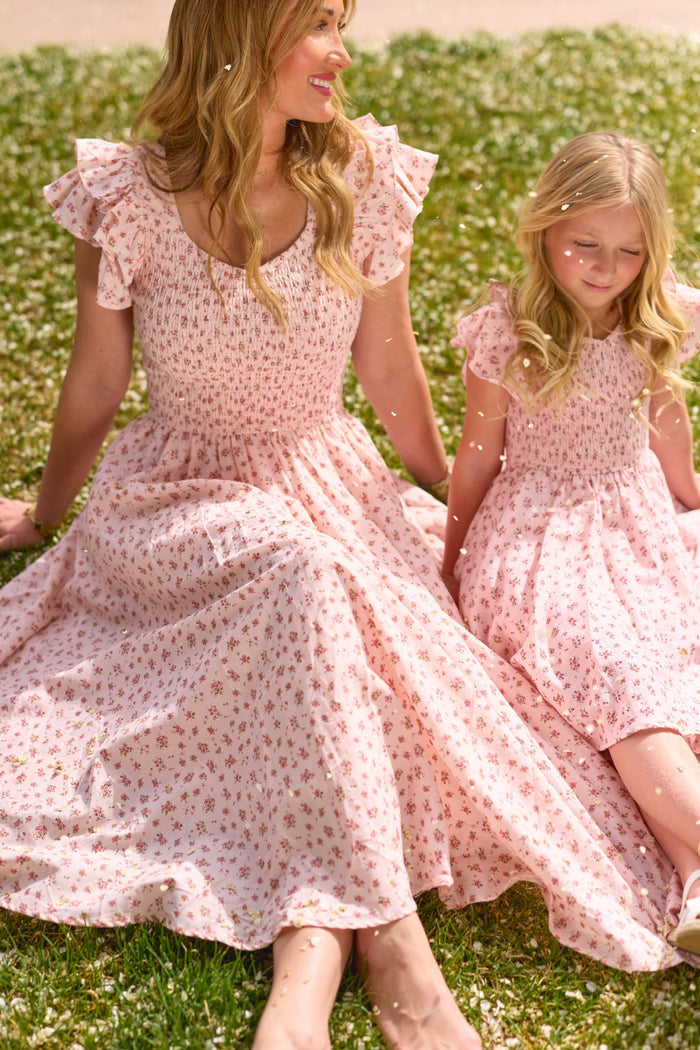 A woman and a young girl, both in Ivy City Co’s Hattie Dress in Madeline Pink, sit on grass with white flowers, smiling and enjoying a sunny day together.