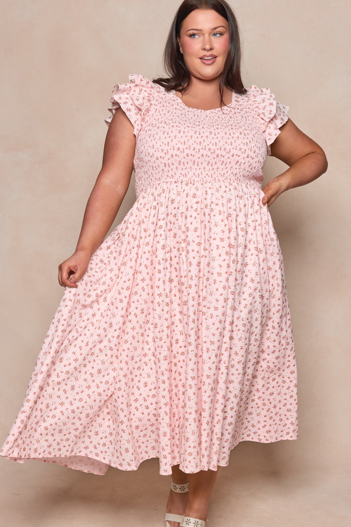 A woman in the Hattie Dress in Madeline Pink by Ivy City Co—a light pink, short-sleeved cotton midi dress with a small floral print—stands against a neutral backdrop, smiling and holding out her dress. A chic choice for a birthday girl.