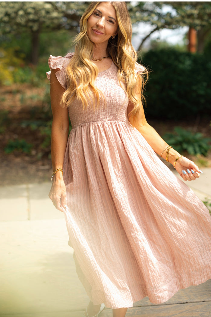 A woman with long, wavy blonde hair stands outdoors in sunlight, smiling softly and holding her skirt while wearing Ivy City Co’s Hattie Dress in Pink Stripes, a beautifully maternity-friendly midi dress with a smocked bodice.