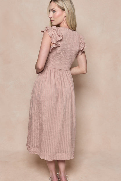 A woman with long blonde hair models the Ivy City Co Hattie Dress in Pink Stripes, featuring a smocked bodice and ruffled sleeves. The maternity-friendly midi is shown from behind as she looks over her shoulder against a beige background.