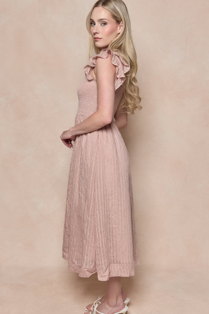 A woman with long blonde hair stands sideways in the Hattie Dress in Pink Stripes by Ivy City Co, featuring ruffle details and a smocked bodice, paired with white sandals against a soft beige background.
