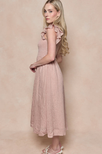 A woman with long blonde hair stands sideways in the Hattie Dress in Pink Stripes by Ivy City Co, featuring ruffle details and a smocked bodice, paired with white sandals against a soft beige background.