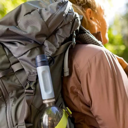 A close-up lifestyle shot of a person wearing a brown hiking jacket and carrying a large olive green backpack outdoors. A LifeStraw Peak Series Solo Water Filter in blue colour is screwed into a water bottle in the side pocket of the backpack.