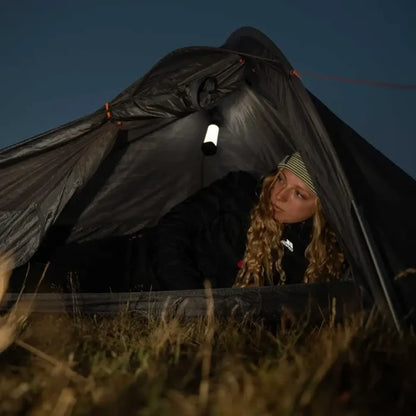 A woman sits inside a dark camping tent at night, illuminated by a Lifesystems Intensity Rechargeable 552 Lumens LED Lantern that is hooked to the roof. The tent is pitched outdoors in grass against a dark blue sky.