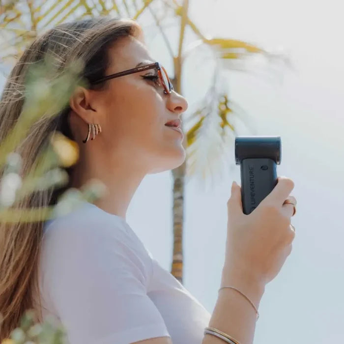 A woman in a white t-shirt and sunglasses holds a Lifeventure Handheld Pocket Fan in profile against a bright outdoor background with palm trees.