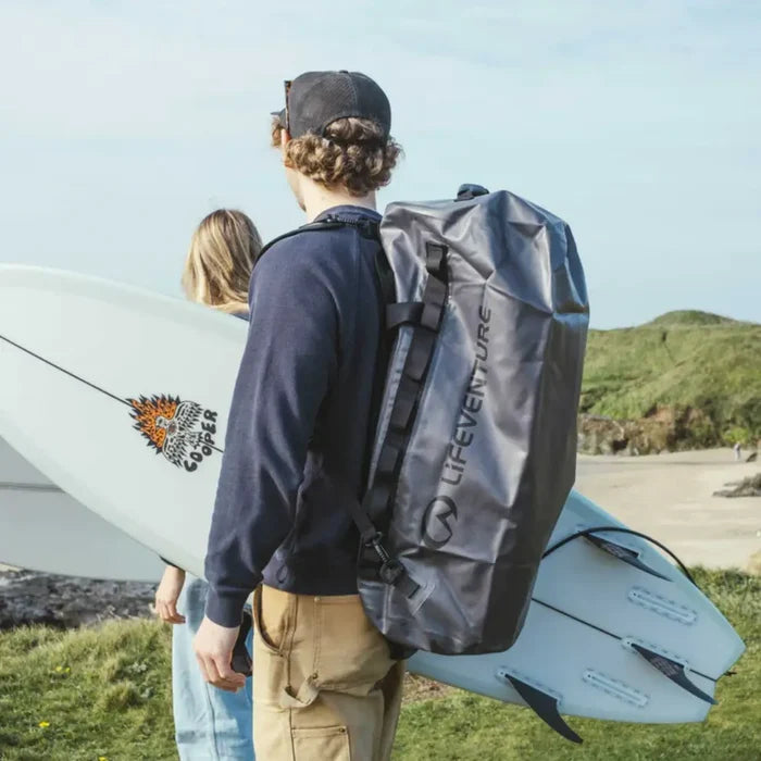 Person carrying a Lifeventure Heavy Duty Dry Bag Duffle 60L on their back with a surfboard on a grassy area.