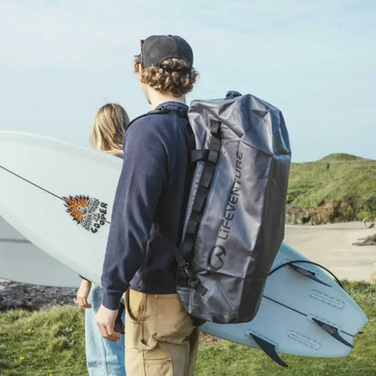Person carrying a Lifeventure Heavy Duty Dry Bag Duffle 60L on their back with a surfboard on a grassy area.