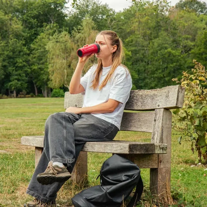 A woman sits on a wooden park bench drinking from a red Lifeventure One Touch Stainless Steel Thermal Mug. She is wearing a white t-shirt, dark gray jeans, and brown hiking shoes, with a black backpack on the grass beside her.