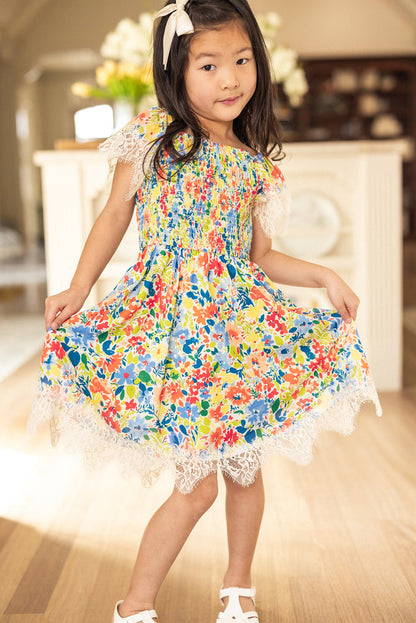 A young girl with long dark hair and a white bow poses indoors in the Ivy City Co Mini Ada Dress in Vibrant Yarrow Print, featuring white lace trim. She wears white sandals and stands on a wooden floor.
