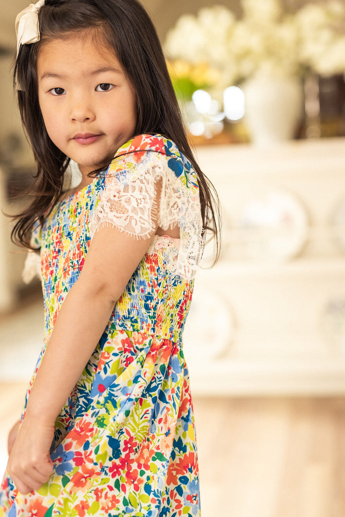 A young girl with long dark hair wears the Mini Ada Dress in Vibrant Yarrow Print by Ivy City Co, standing indoors in a bright room with a soft background of flowers and decorative plates.